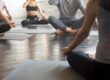 Group of people sitting cross-legged on yoga mats in a studio, practicing meditation with hands resting on knees in a calm, relaxed posture.