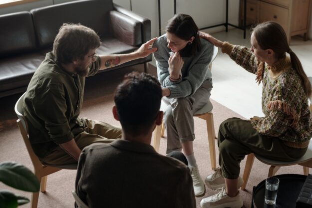A woman sits with her head lowered in a support group as three other members gently touch her shoulders and back, offering comfort and encouragement.