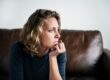 A woman with short curly blonde hair sits on a brown leather couch, resting her chin on her hand and gazing thoughtfully to the side.