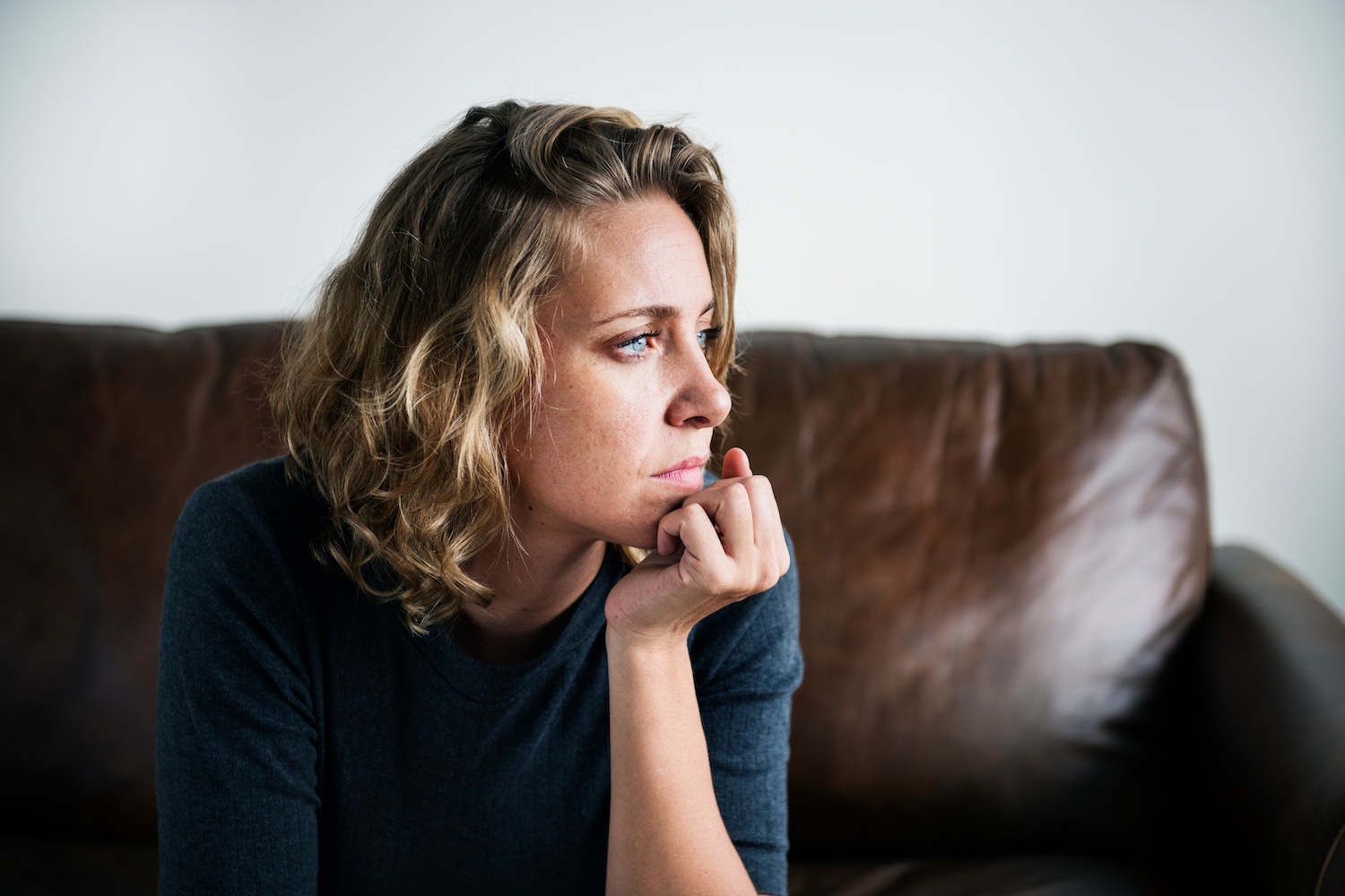 A woman with short curly blonde hair sits on a brown leather couch, resting her chin on her hand and gazing thoughtfully to the side.