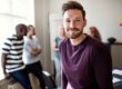 Confident young man smiling at the camera in an office setting, with a group of colleagues talking and collaborating in the background.