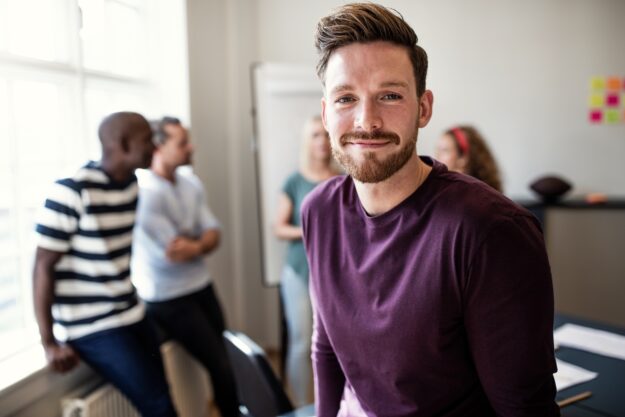 Confident young man smiling at the camera in an office setting, with a group of colleagues talking and collaborating in the background.