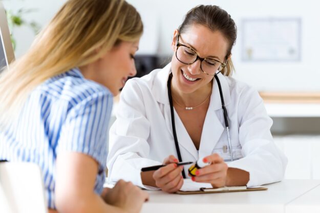 Smiling doctor wearing a white coat and stethoscope showing a medication bottle to a patient during a consultation at a medical office.