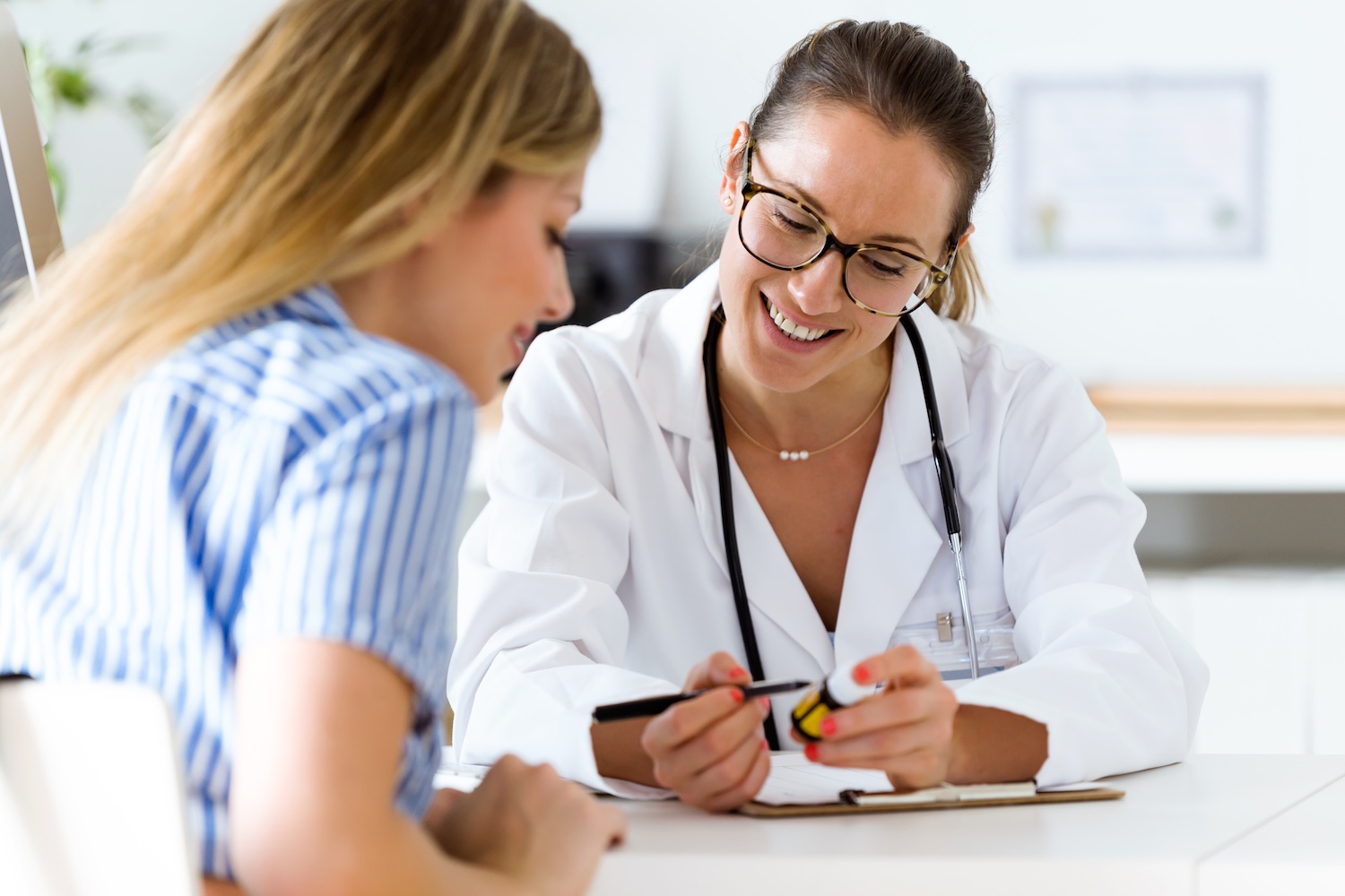Smiling doctor wearing a white coat and stethoscope showing a medication bottle to a patient during a consultation at a medical office.
