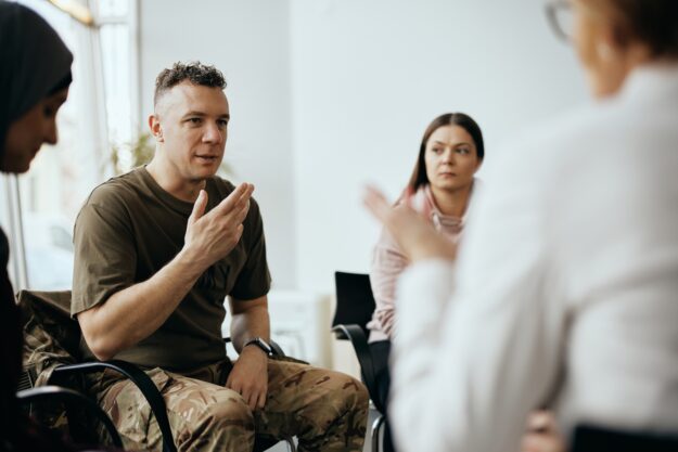 A man in military-style clothing speaks during a group therapy session, gesturing with his hand while other participants listen attentively.