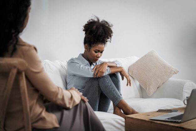 A young woman sits curled up on a couch with her head down and arms wrapped around her knees, looking distressed, while another woman sits nearby, watching with concern.