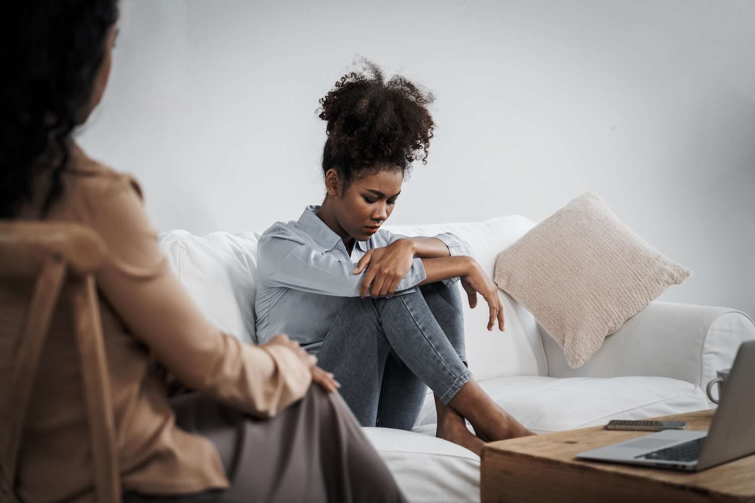 A young woman sits curled up on a couch with her head down and arms wrapped around her knees, looking distressed, while another woman sits nearby, watching with concern.