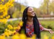 Woman standing outdoors in a park surrounded by bright yellow flowers, smiling with her eyes closed and arms outstretched, enjoying the sunshine and nature.