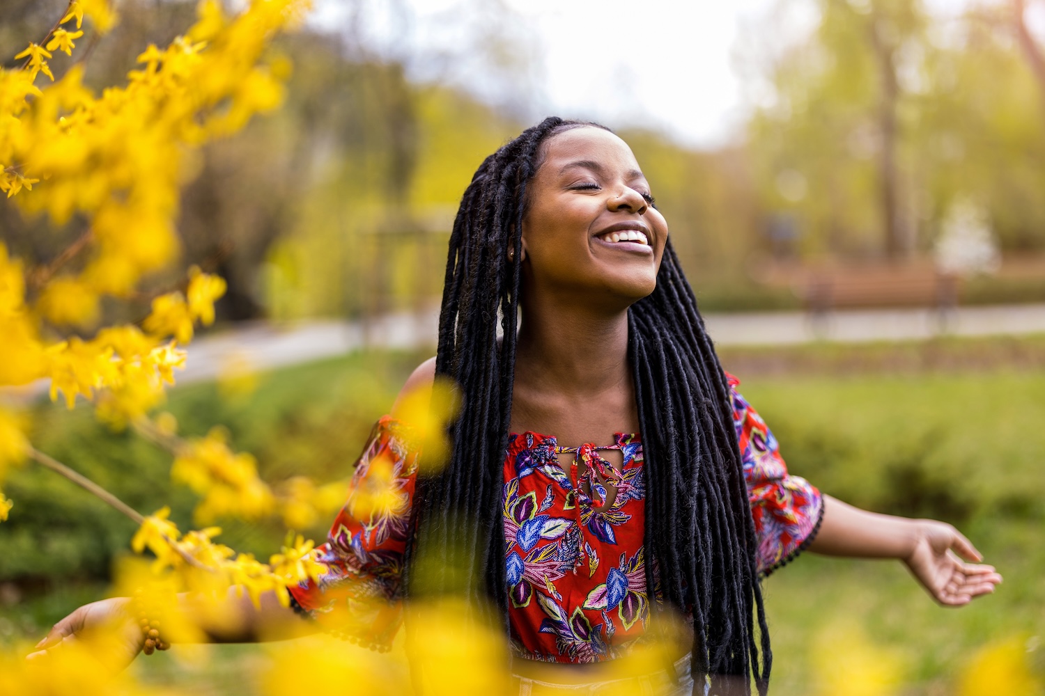Woman standing outdoors in a park surrounded by bright yellow flowers, smiling with her eyes closed and arms outstretched, enjoying the sunshine and nature.