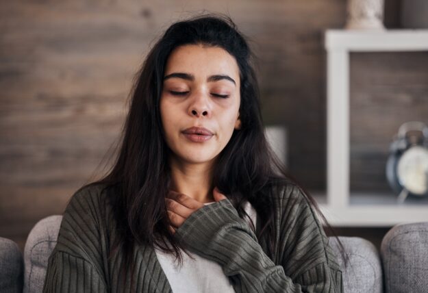 A woman with long dark hair sits indoors with her eyes closed, one hand resting on her chest, appearing to take a deep breath or calm herself, with a neutral-toned room in the background.