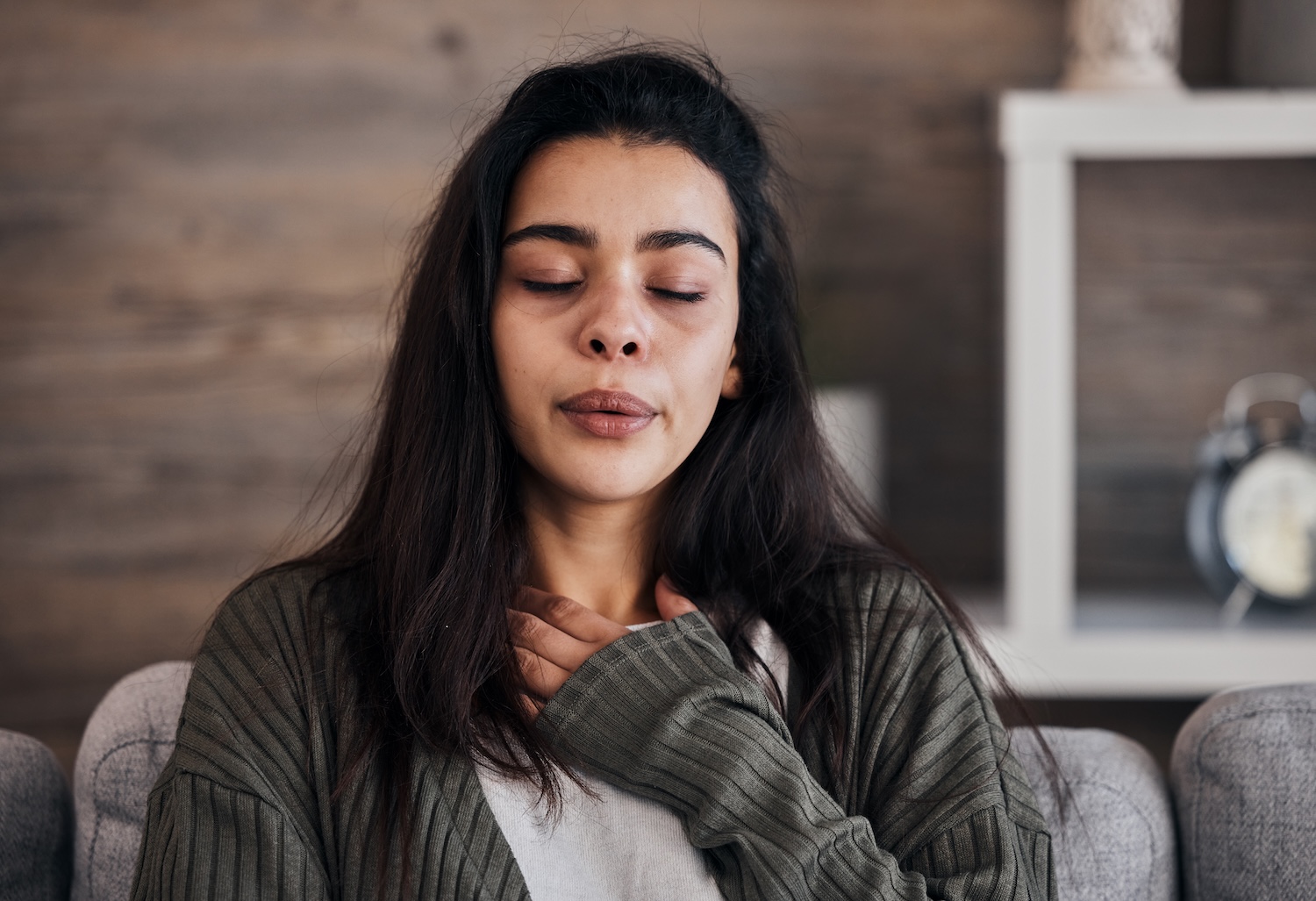 A woman with long dark hair sits indoors with her eyes closed, one hand resting on her chest, appearing to take a deep breath or calm herself, with a neutral-toned room in the background.