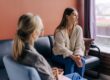 A woman sits on a couch looking out the window with a thoughtful, slightly concerned expression while another woman, possibly a therapist, sits across from her listening attentively.
