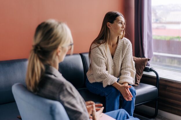 A woman sits on a couch looking out the window with a thoughtful, slightly concerned expression while another woman, possibly a therapist, sits across from her listening attentively.