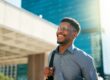 A smiling man wearing glasses and a button-up shirt walks outdoors in a city, carrying a backpack over one shoulder with tall modern buildings in the background.