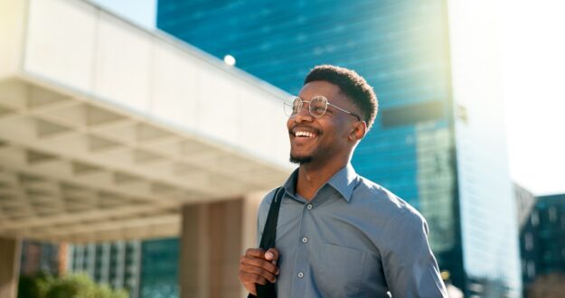 The Benefits of Combining Detox with Therapy for Sustainable Recovery A smiling man wearing glasses and a button-up shirt walks outdoors in a city, carrying a backpack over one shoulder with tall modern buildings in the background.