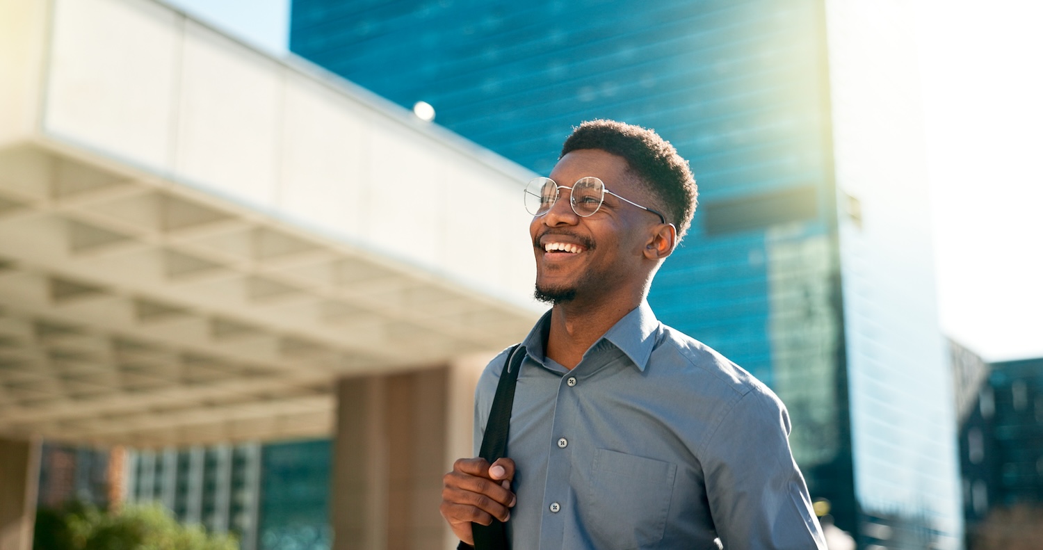 A smiling man wearing glasses and a button-up shirt walks outdoors in a city, carrying a backpack over one shoulder with tall modern buildings in the background.