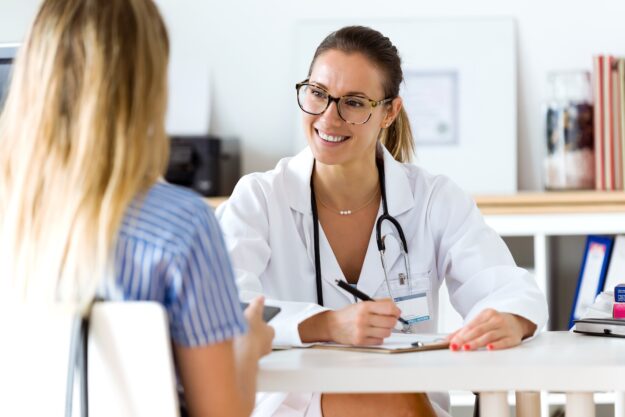 A smiling female doctor wearing glasses and a white coat sits at a desk with a clipboard, talking to a patient whose back is to the camera in a bright medical office.