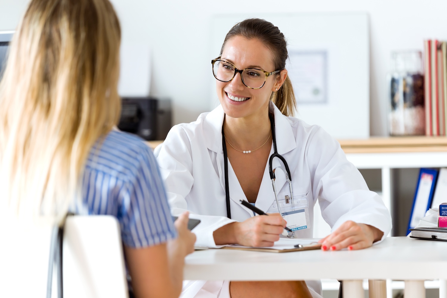 A smiling female doctor wearing glasses and a white coat sits at a desk with a clipboard, talking to a patient whose back is to the camera in a bright medical office.